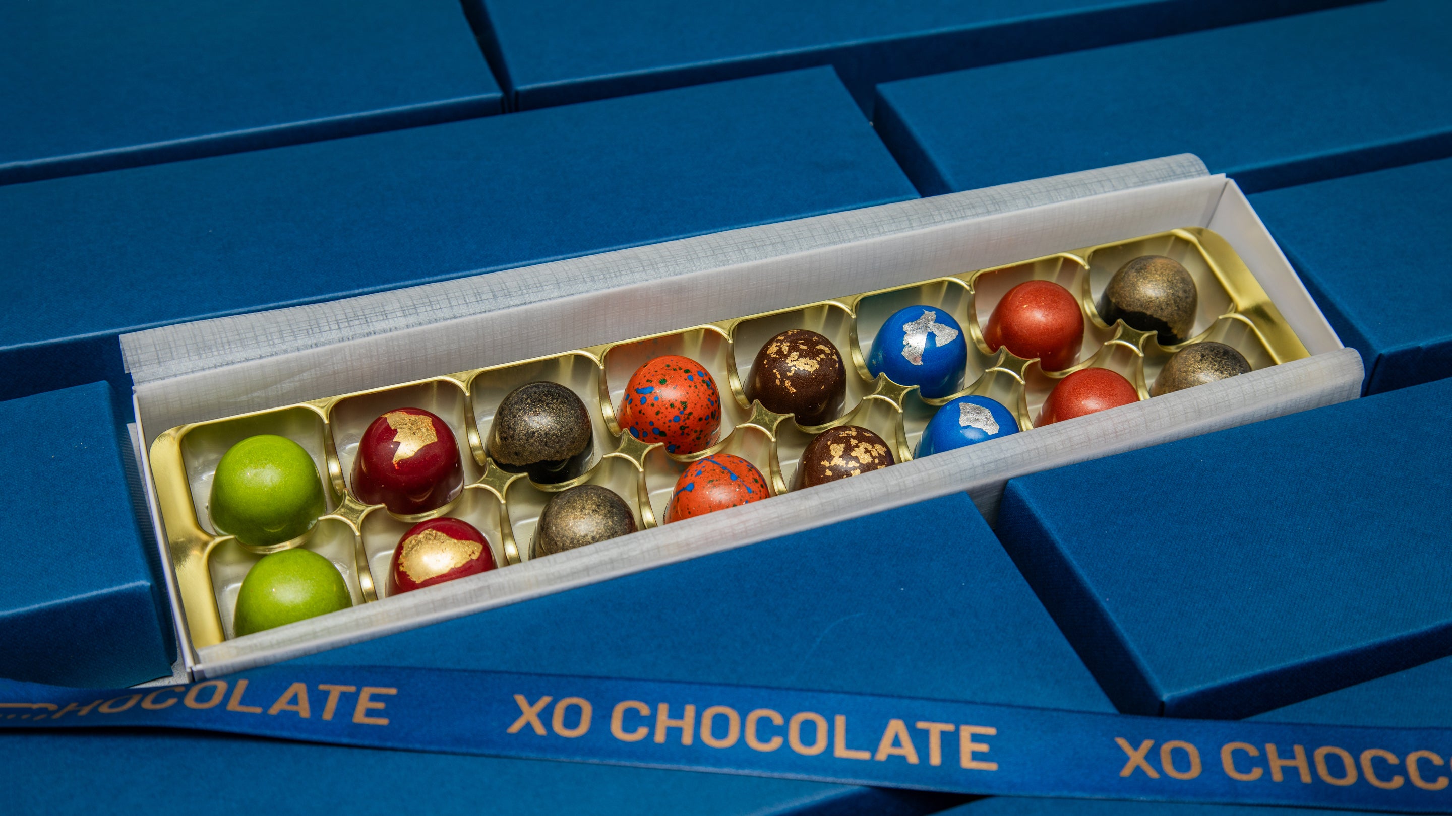 An open box of XO Chocolate truffles displayed diagonally on a blue background. The box contains 12 assorted chocolates covered in various colors including green, red, blue, brown, and gold.