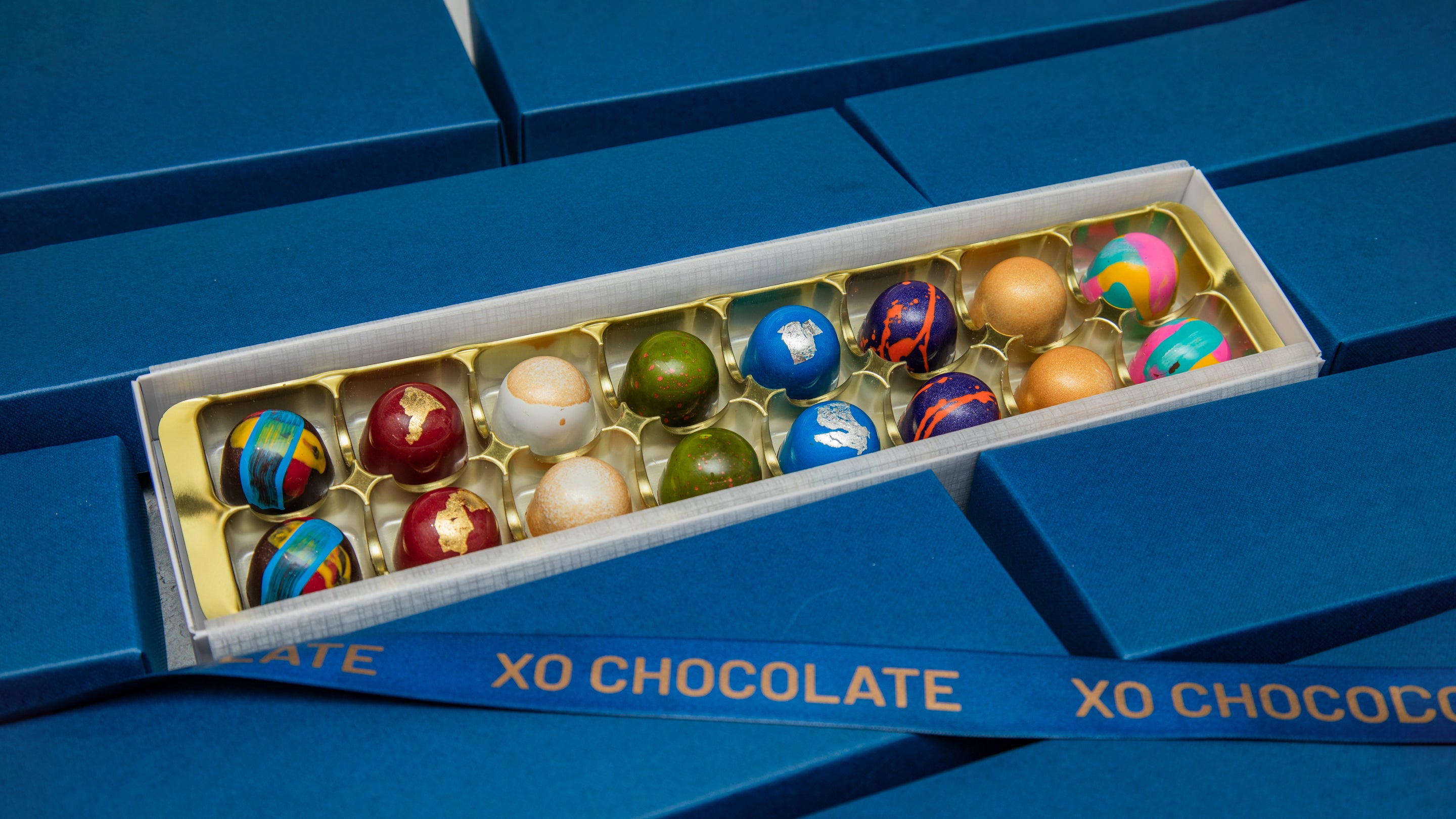 An open box of XO Chocolate truffles displayed on a blue background. The box contains assorted chocolate truffles in colorful, patterned milk and dark chocolate shells, including blue, green, red, and gold designs.
