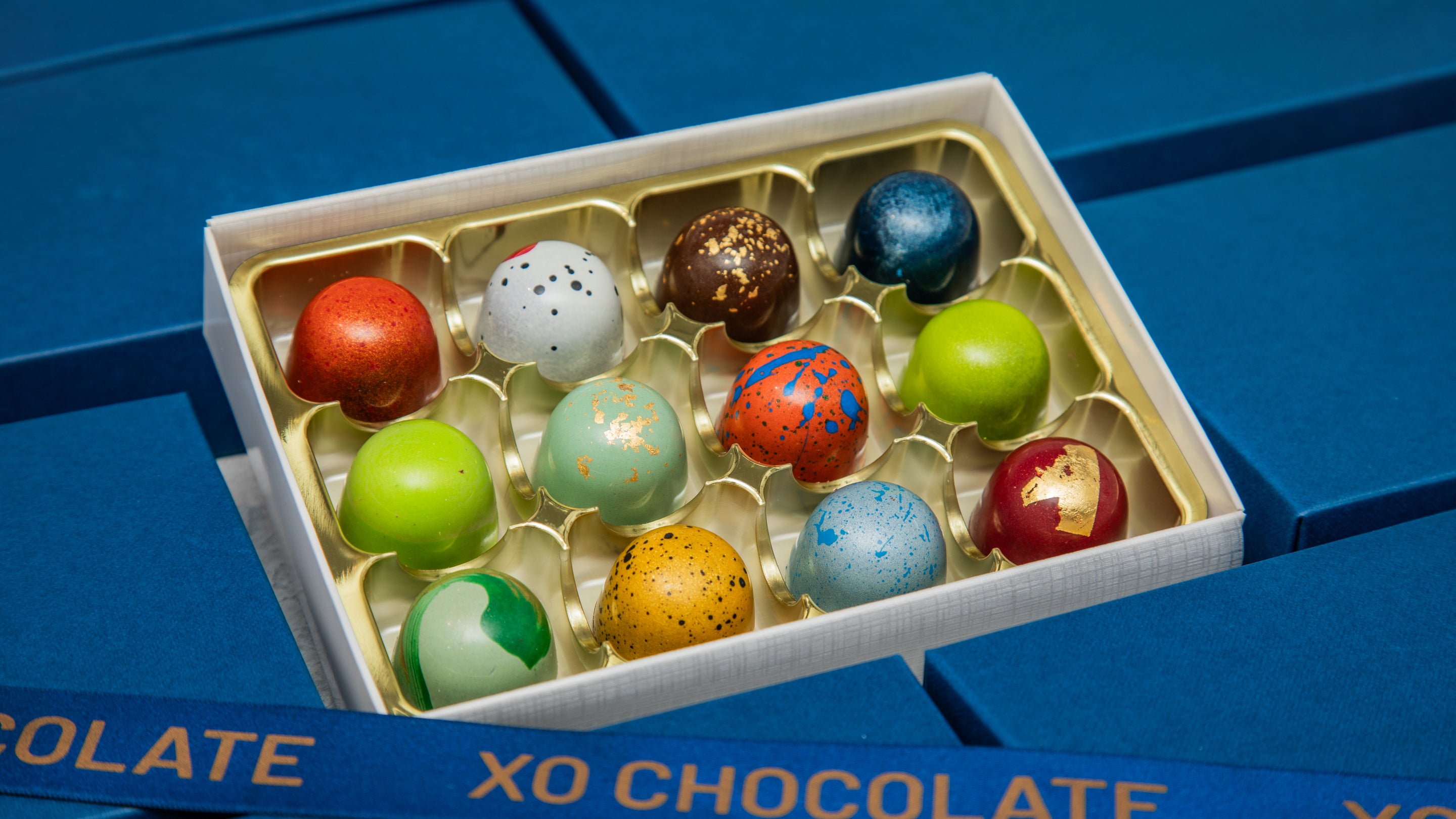 An open box of assorted chocolate truffles with colorful, decorated chocolates arranged in a white and gold tray, set against a blue chocolate box background.