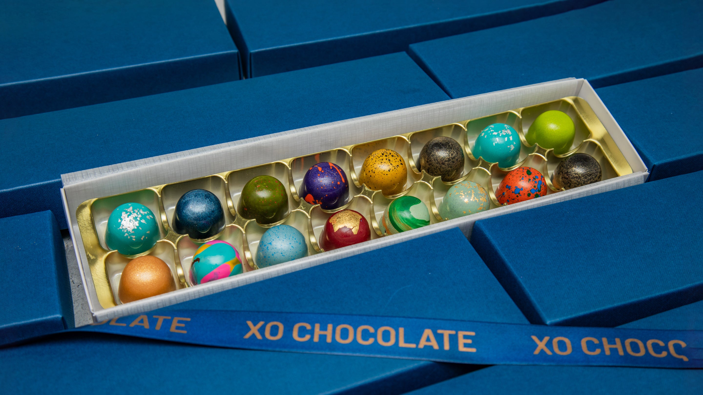 An open box of XO Chocolate truffles displayed on a blue background. The box contains 18 individual chocolate truffles in colorful, decorative design.