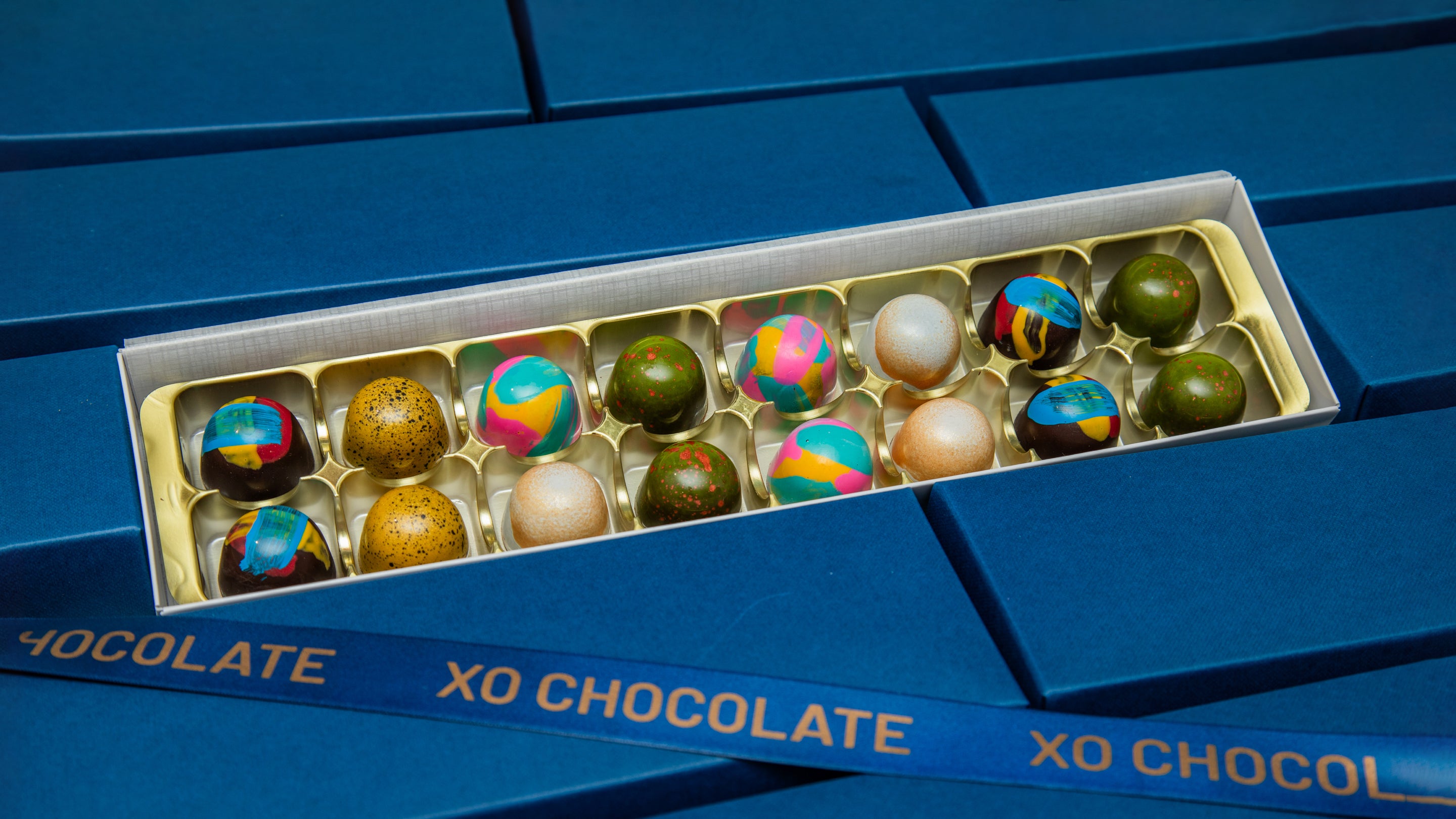 An open box of XO Chocolate truffles displayed on a blue textured background. The box contains 12 assorted chocolates, including green, brown, and colorful patterned varieties, with the brand name 'XO CHOCOLATE' printed on the blue lid.
