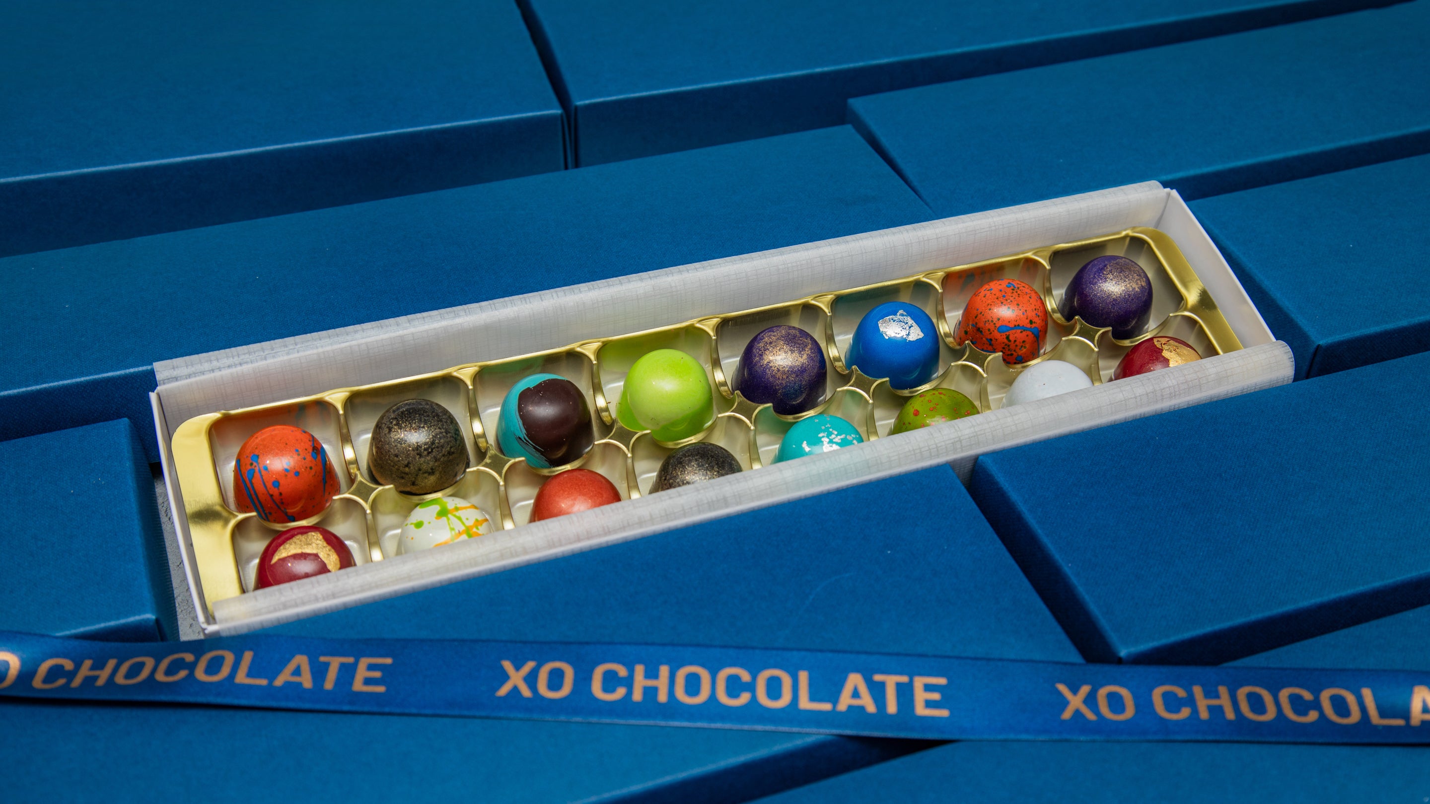 An open box of XO Chocolate truffles displayed on top of a blue box. The box contains assorted round chocolate truffles in various colors, including red, green, blue, purple, and brown, set against a blue background.