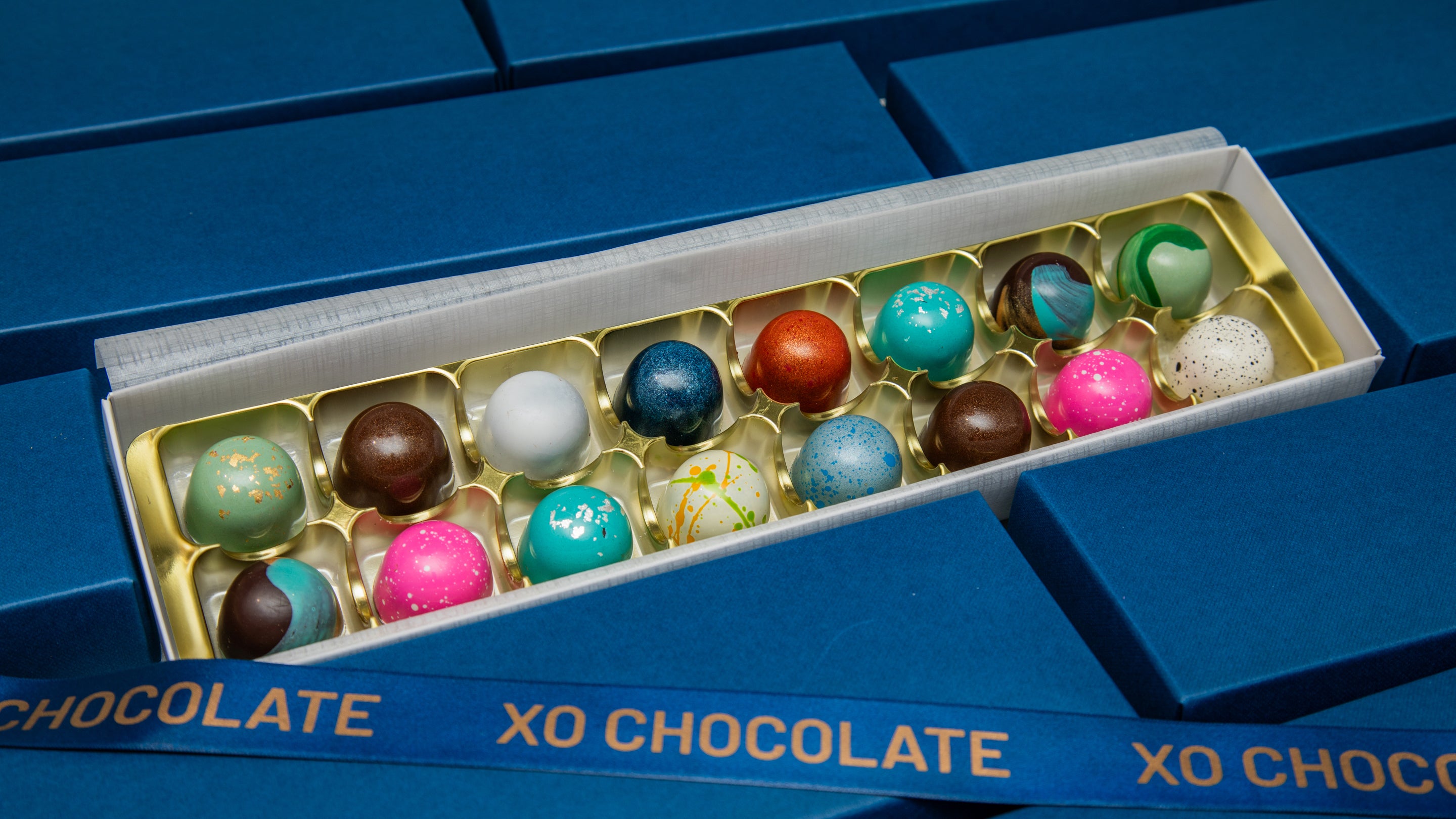 An open box of XO Chocolate truffles displayed diagonally on a blue chocolate box. The box contains 16 assorted chocolate-covered spheres in various colors, including blue, pink, green, brown, and white, set in a gold tray.